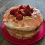 A 2 layer sponge cake with buttercream and roasted Strawberries on the top. In between thr layers of cake more buttercream icing is visible with hints of strawberry juice oozing onto the sponge. It is plated on a vibrant pink plate