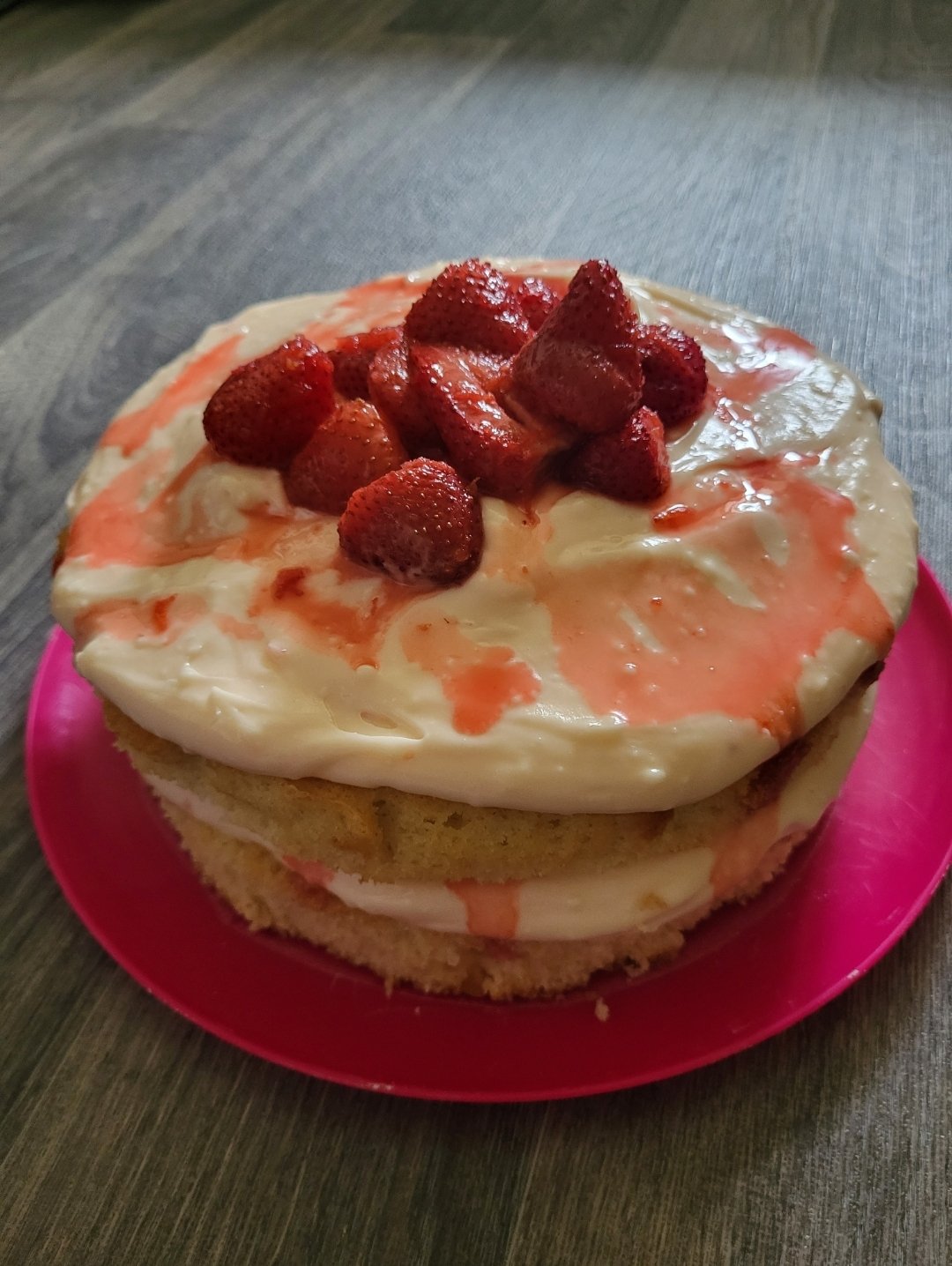 A 2 layer sponge cake with buttercream and roasted Strawberries on the top. In between thr layers of cake more buttercream icing is visible with hints of strawberry juice oozing onto the sponge. It is plated on a vibrant pink plate