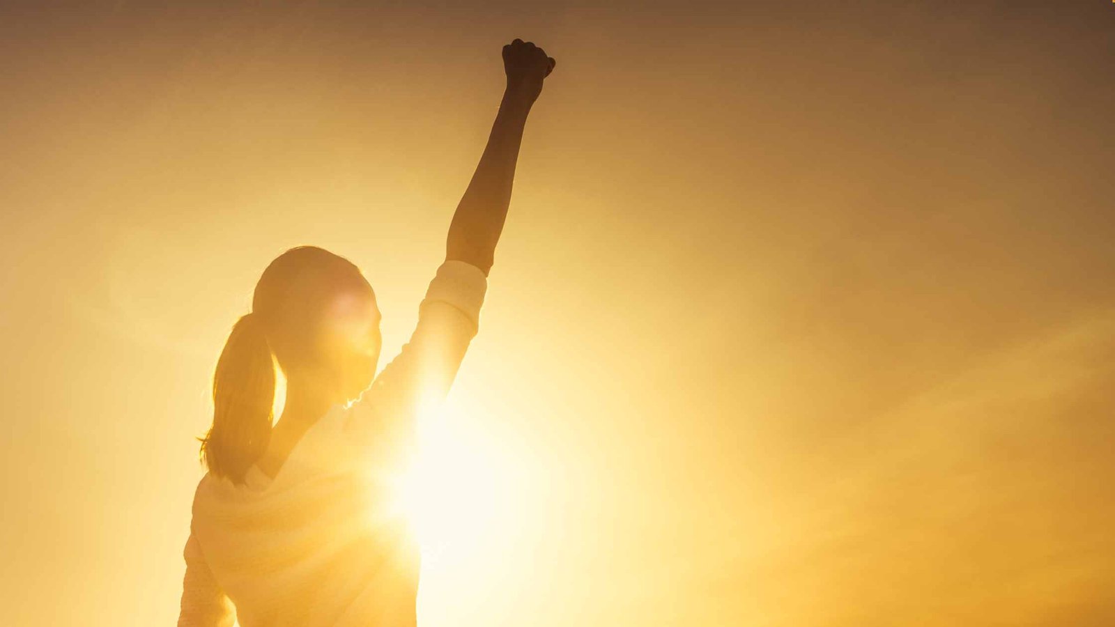 A silhouette of a woman with long hair stood in front of the sun rising with one arm up above her head in triumph