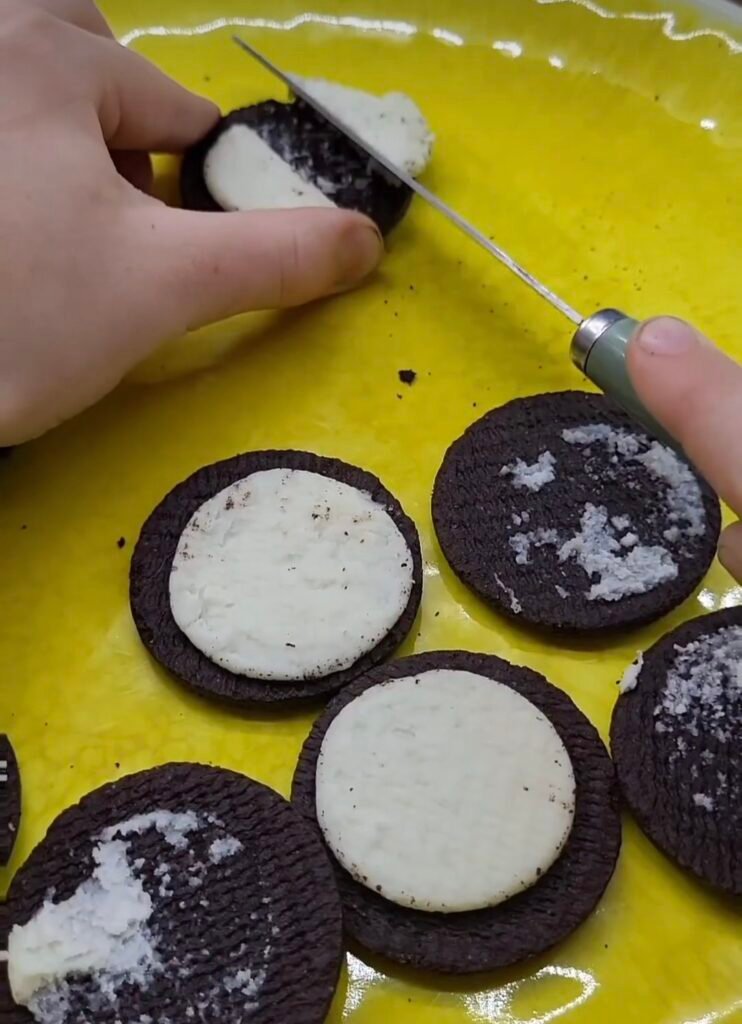 A child's hand holds an oreo  and using a butter knife is scraping half of the cream from the biscuit