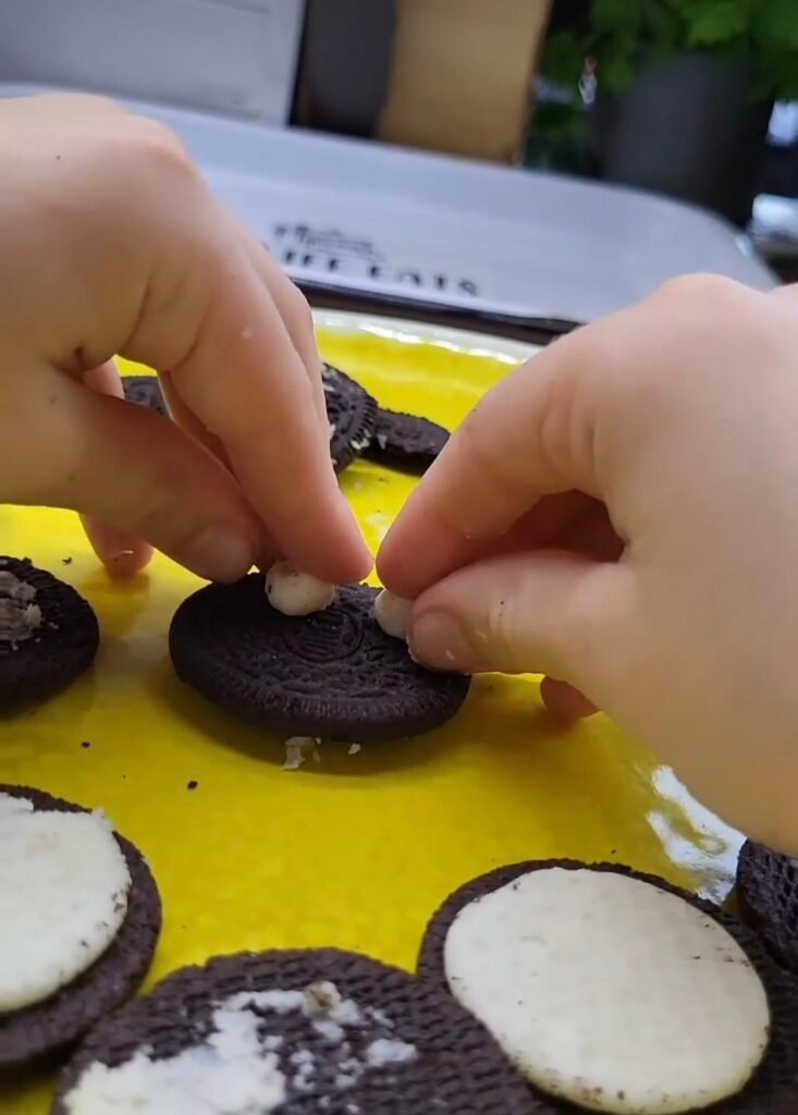 A child's hands placed 2 rolled up balls of white icing onto an oreo