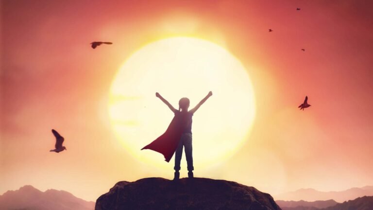 The photo shows a child from behind, They are standing on a large rock with the sun setting in front of them so they are a silhouette. Birds can be seen flying in the red coloured sun. The child has both arms above their head and a superhero cape is billowing out behind them