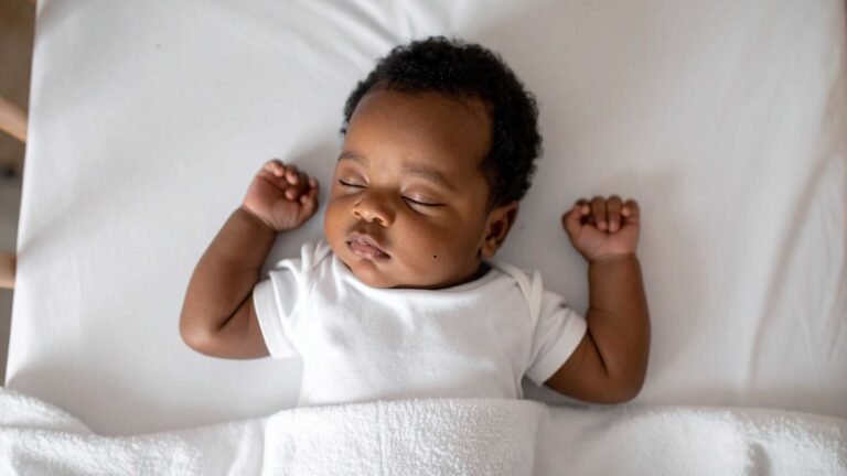The photo shows a baby asleep, with a white sheet behind them, they are wearing a white baby vest and a white blanket is covering them. Both their fists are balled with their arms up by their head